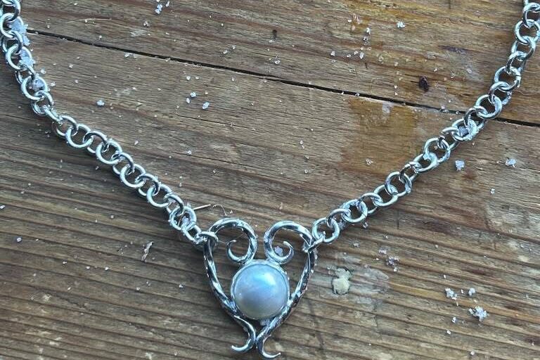 A silver chain necklace with a pearl in a scrolled setting, lying on a wooden workbench